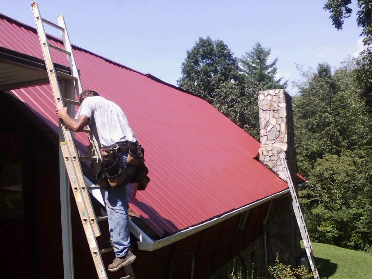 Roofer on a ladder beside a red metal roof and stone chimney — Soffit & Fascia Repair by Roofing Brothers in Cookstown